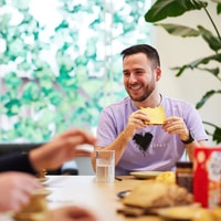 Man geniet van ontbijt met kaas, glimlachend aan tafel in een lichte, groene kantoorruimte. Ontspannen sfeer.