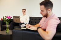 Man met laptop glimlacht, zittend op een bank met een andere man die notities maakt, bloemen in de voorgrond.