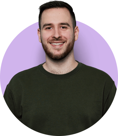 Young man smiling in a dark green crewneck sweater, pictured against a light purple background.