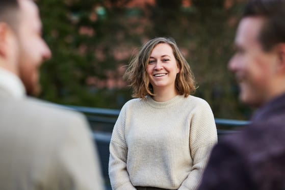Persoon glimlacht naar collega’s tijdens een informeel gesprek buiten op een zonnige dag.