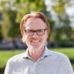Smiling man with glasses and red hair standing outside in a park-like setting on a sunny day.