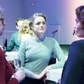 Jongere vrouw in een blauwe trui in gesprek met anderen tijdens een netwerkevenement, omringd door zacht licht.