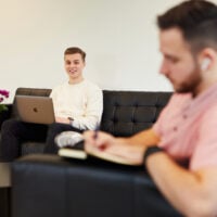 Man met laptop glimlacht, zittend op een bank met een andere man die notities maakt, bloemen in de voorgrond.