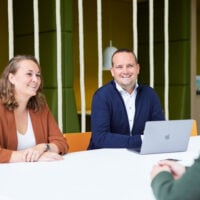 Twee mensen aan tafel met laptop tijdens een collegiale vergadering in een moderne kantooromgeving.