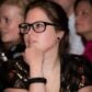 Young woman with glasses pondering, wearing black sequined top, in a crowd at an event.