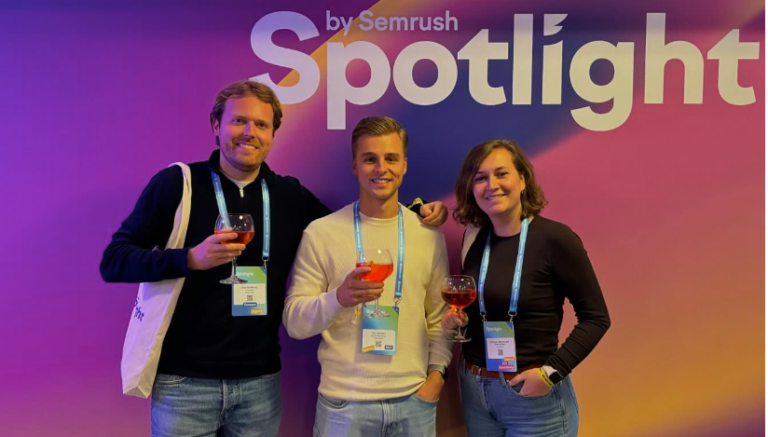 Drie mensen poseren voor een Semrush Spotlight-bord met drankjes, lachend en dragen conferentie-badges.